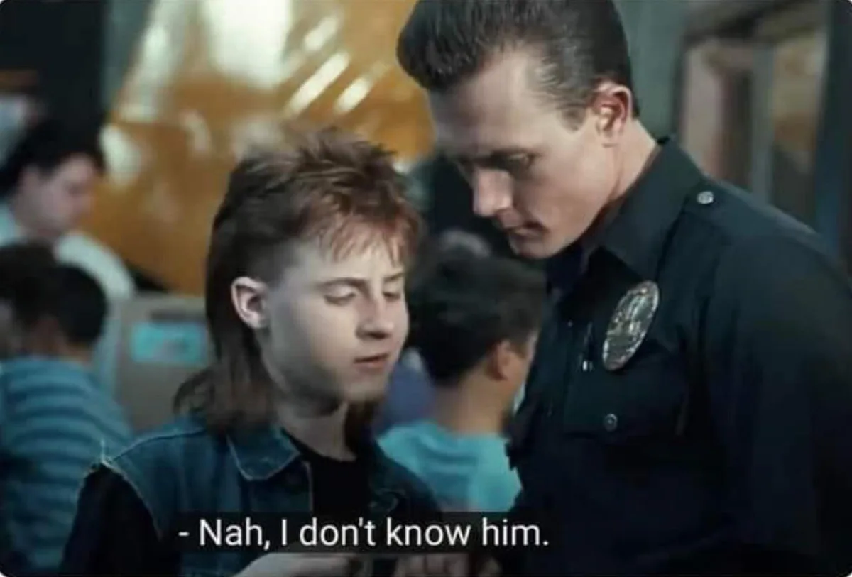 A young man with a mullet and denim vest looks down while a police officer leans in to question him. The officer has a stern…
