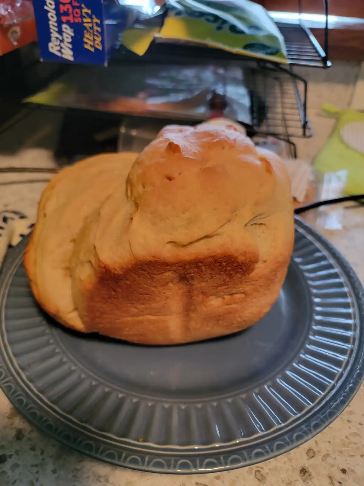 A golden-brown, round loaf of bread sits on a dark blue glass plate on a kitchen counter. In the background, a box of…