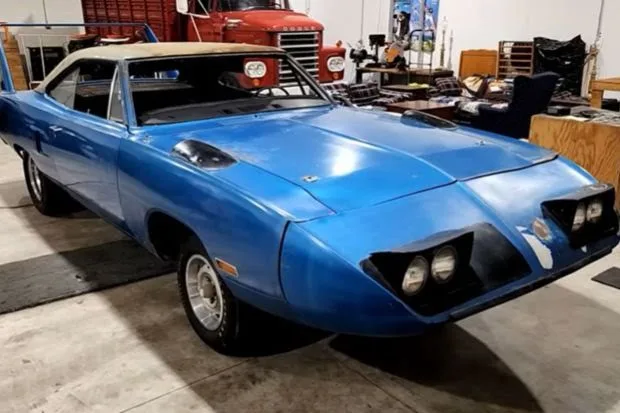 a blue 1970 dodge challenger t/A with a custom hood scoop and tan vinyl top is parked in a cluttered garage filled with…