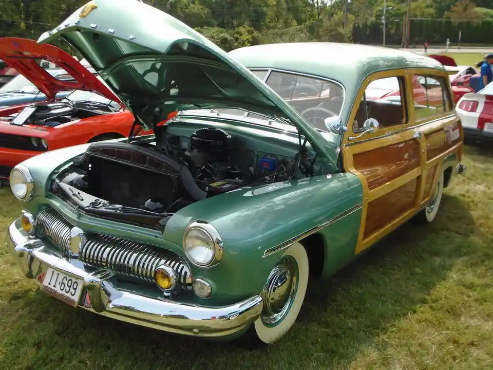 A vintage green and wood-paneled station wagon with its hood open, parked on grass at a car show. Other classic cars are…