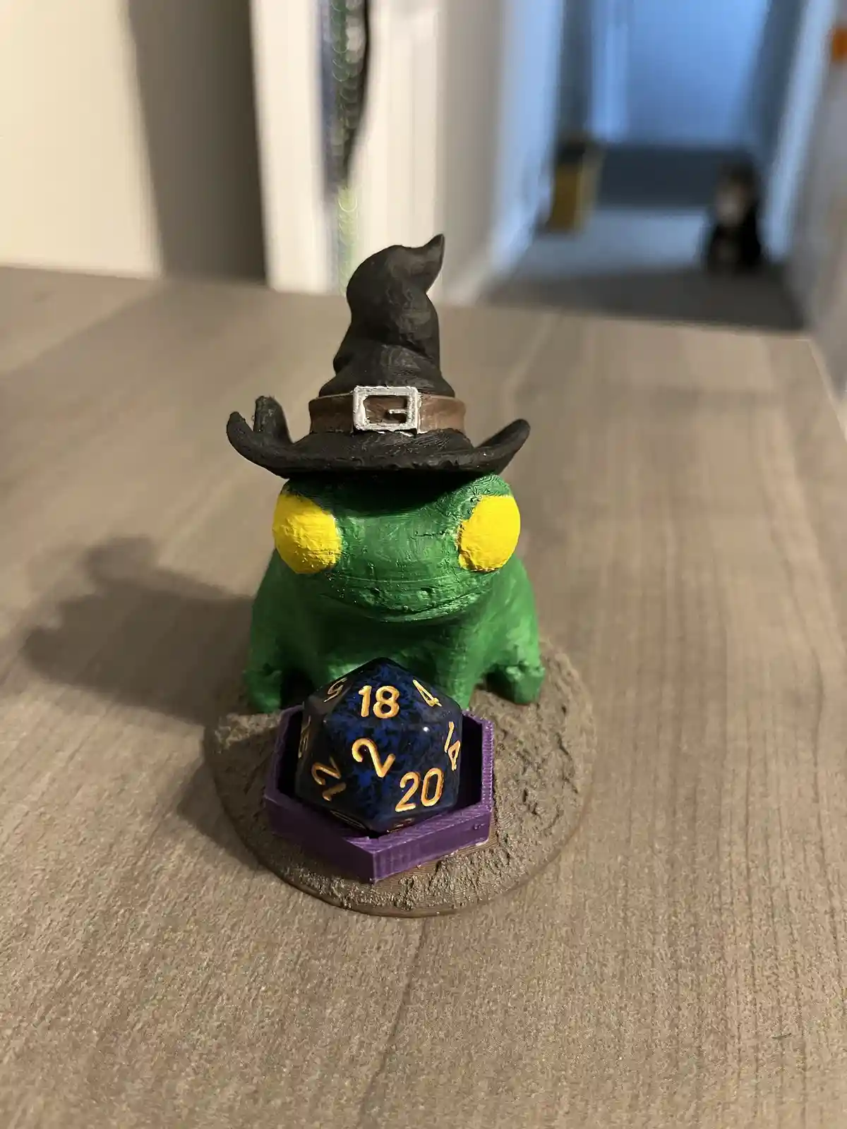 A green, frog-like figurine wearing a black witch's hat with a silver buckle sits on a textured base. In front of it, a blue…