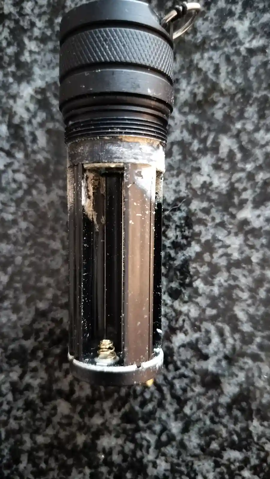 A close-up photo of a black tactical flashlight with its lower half visibly corroded and covered in white residue, sitting…