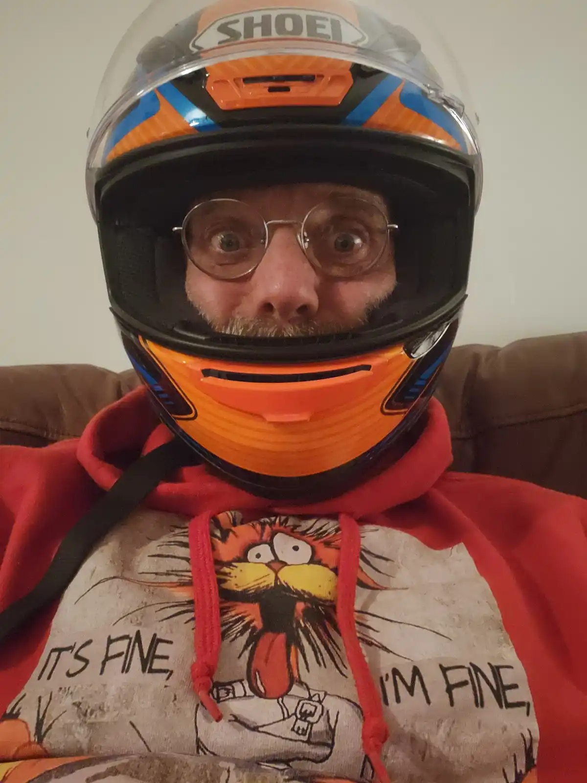A man with round glasses and a mustache wears a full-face orange and blue Shoei motorcycle helmet while sitting on a brown…