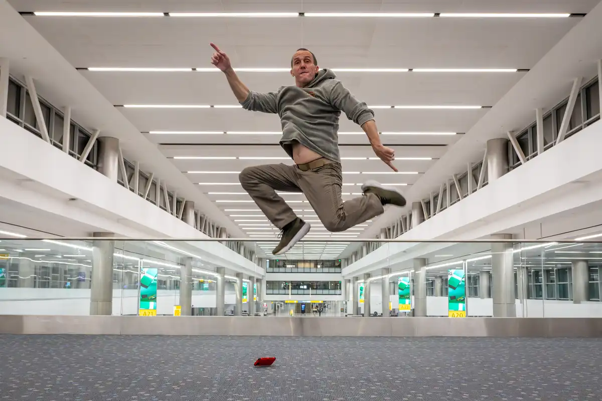 A man is captured mid-air, jumping enthusiastically in a modern airport terminal with glass railings and overhead lighting…