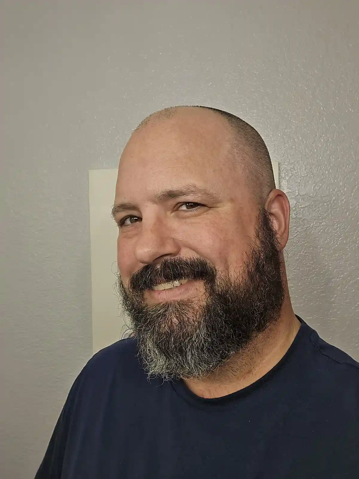 A smiling man with a shaved head and a full beard, wearing a dark blue t-shirt, stands against a textured light-colored…