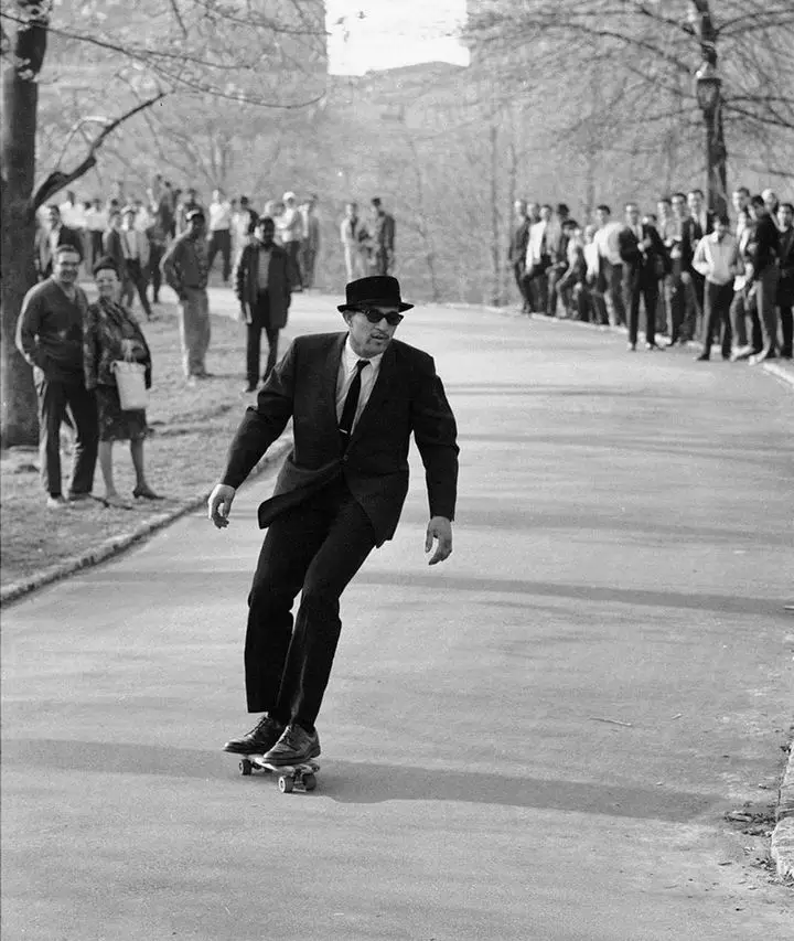 a black-and-white photo shows a man in a suit, fedora, and sunglasses riding a skateboard down a paved path in a park, with…