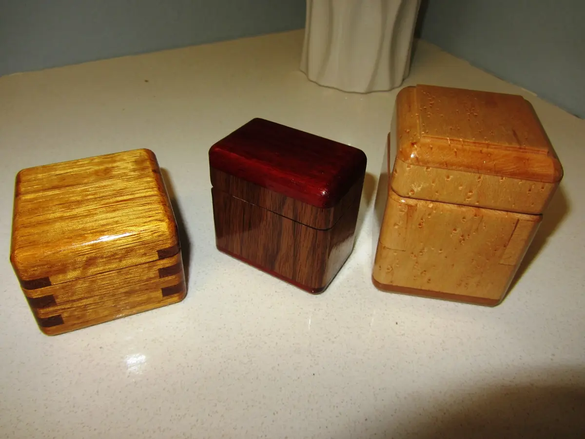 Three handcrafted wooden boxes of varying wood types and finishes sit on a white countertop. The left box has a light wood…