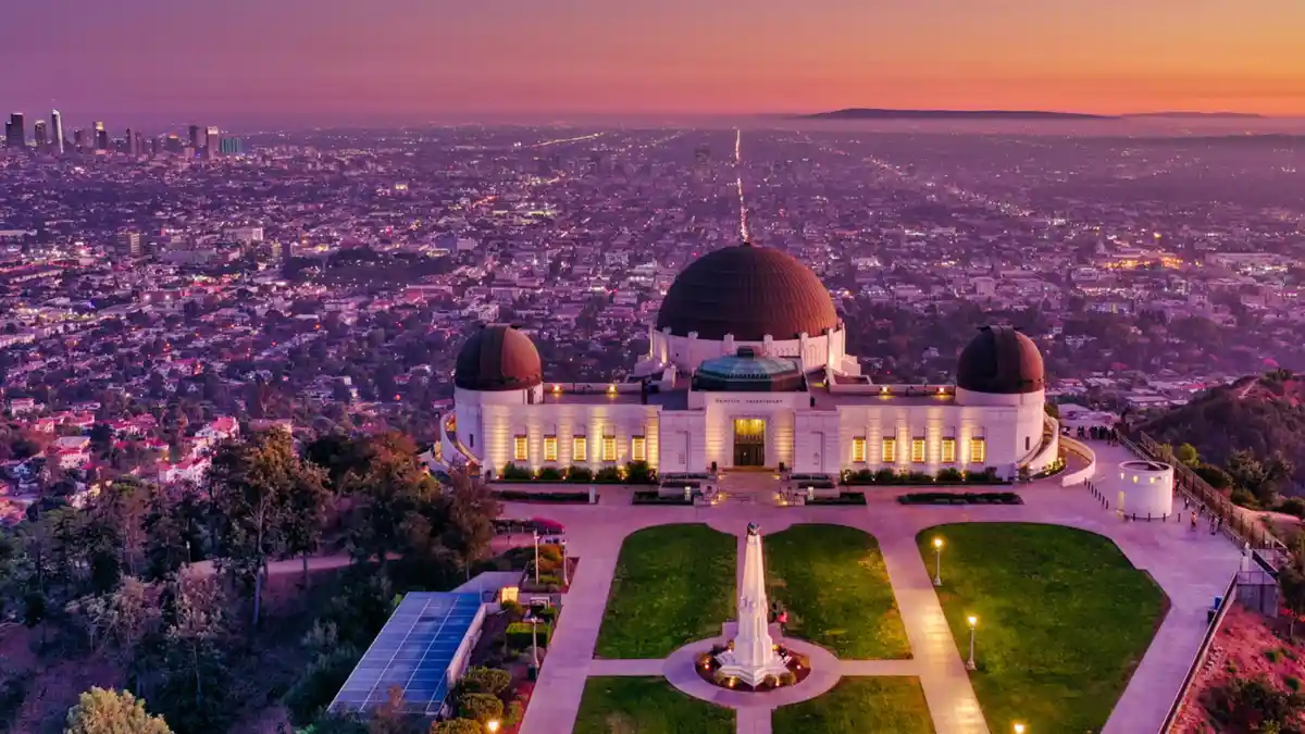 An aerial view of the Griffith Observatory at dusk, illuminated against a purple and orange sky, overlooking the sprawling…