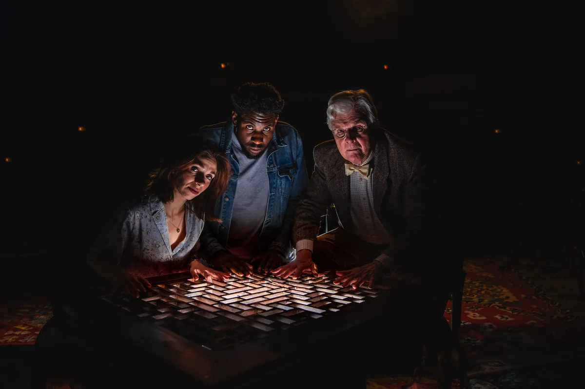 Three people lean over a glowing, grid-like table in a dark room, all staring intently with expressions of confusion and…