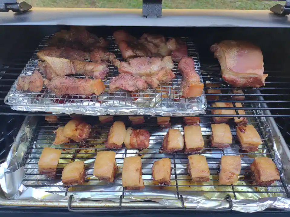 A close-up photo of a two-tiered grill filled with various cuts of meat, including bacon strips, pork belly, and larger…