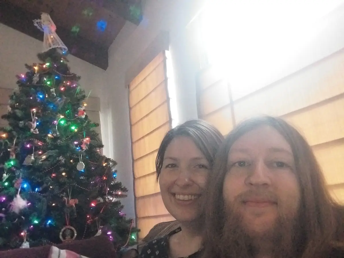 A selfie of two people smiling in front of a brightly lit, decorated Christmas tree with colorful lights and ornaments. The…