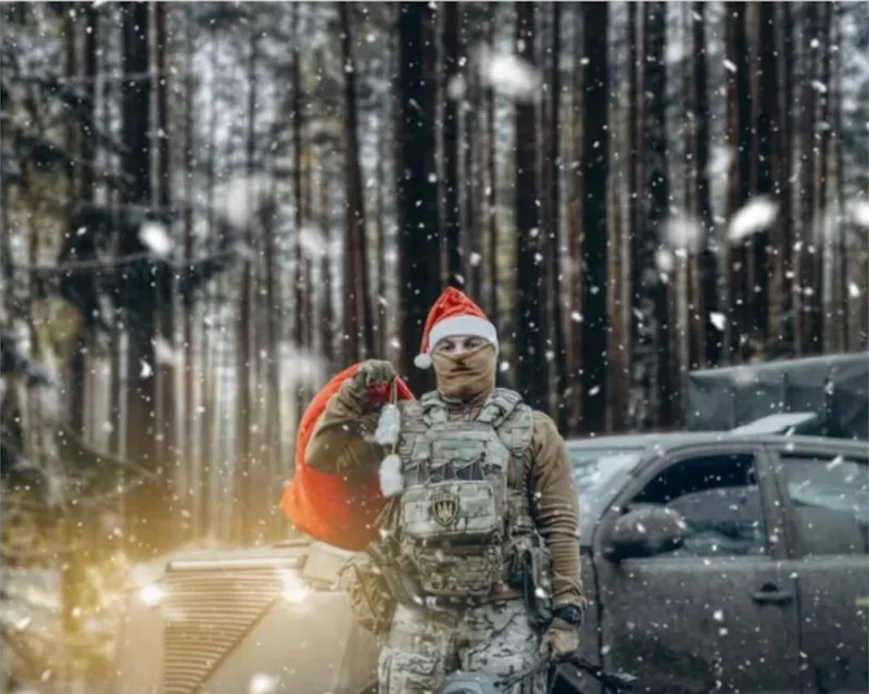 a soldier in full tactical gear and a santa hat stands in a snowy forest, holding a red santa sack, with a military vehicle…