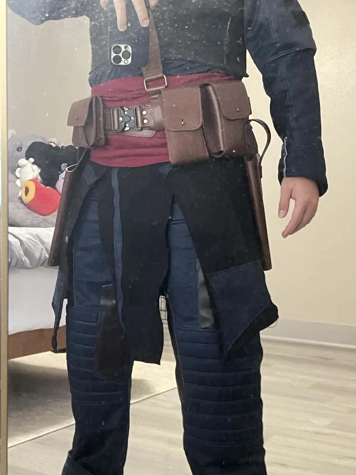 A person in a Star Wars-inspired costume with a brown leather belt, pouches, and padded pants stands in front of a mirror…