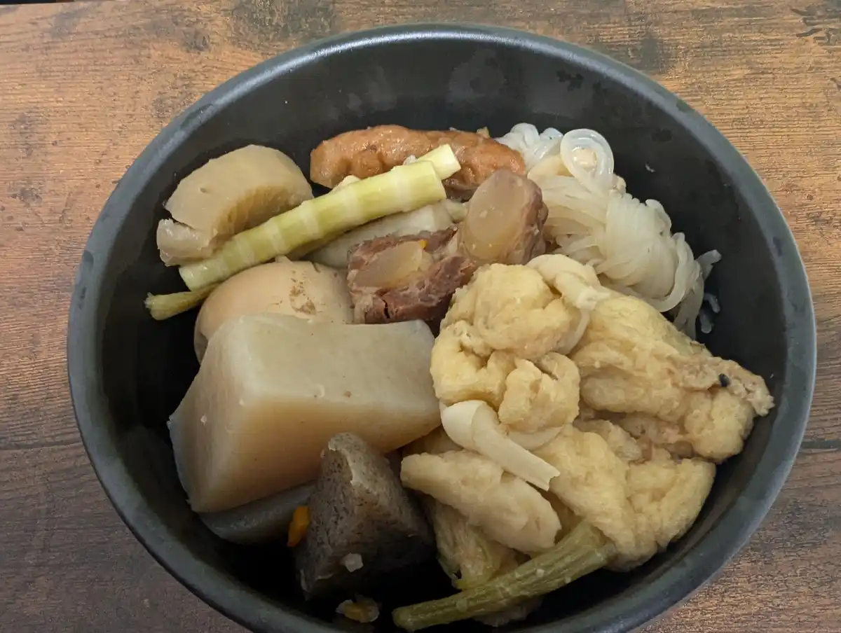 a black bowl filled with various cooked ingredients including noodles, tofu, meat, bamboo shoots, and other vegetables sits…