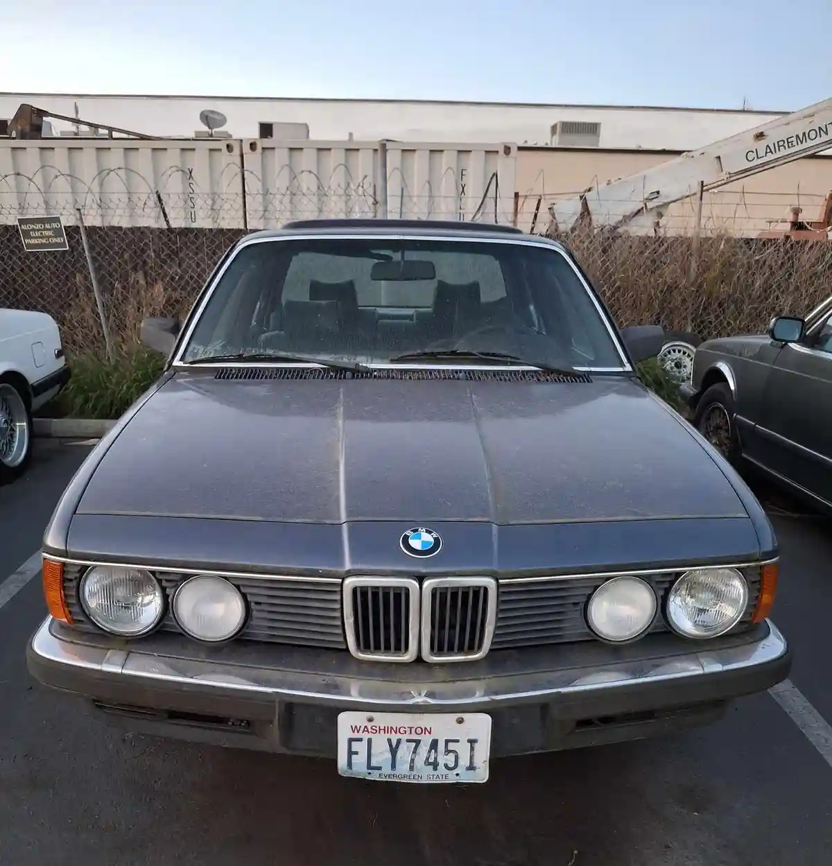 A dusty, dark gray BMW sedan with a Washington license plate reading 'FLY745I' is parked in a lot. The car has a classic…