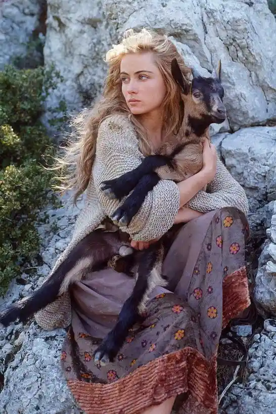 a woman with long blonde hair sits on rocky terrain holding a small black and tan goat. She wears a beige knit sweater and a…