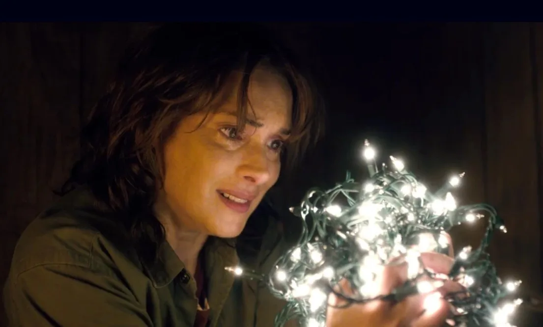 A woman with a tear-streaked face and a sad, pained expression holds a tangled string of bright white Christmas lights in…