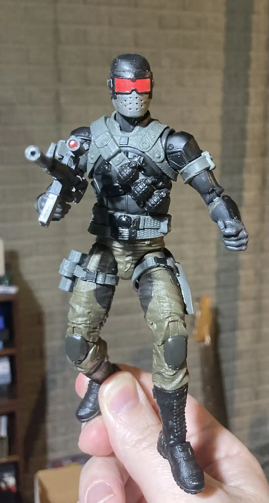 A person holds a detailed action figure of a soldier with red visor goggles, tactical armor, and a weapon. The figure is…