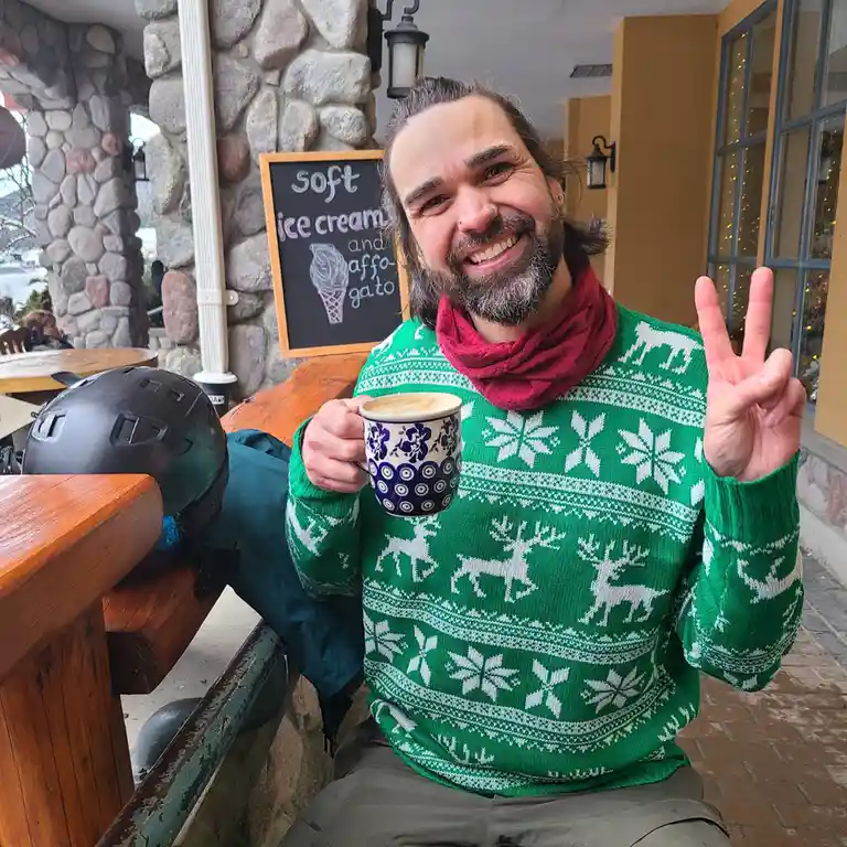 A man with a beard and long hair, wearing a green holiday sweater with reindeer and snowflakes, sits outside a cafe holding…