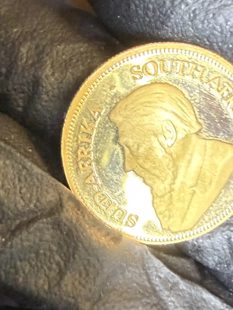 A close-up photo shows a gold-colored coin with the profile of a man and the words 'SOUTH AFRICA' inscribed along its edge…