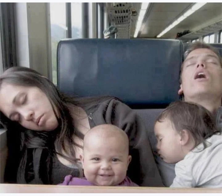 A family of four is asleep on a train seat: a woman slumped over, a man with his mouth open, and two babies. The baby in the…