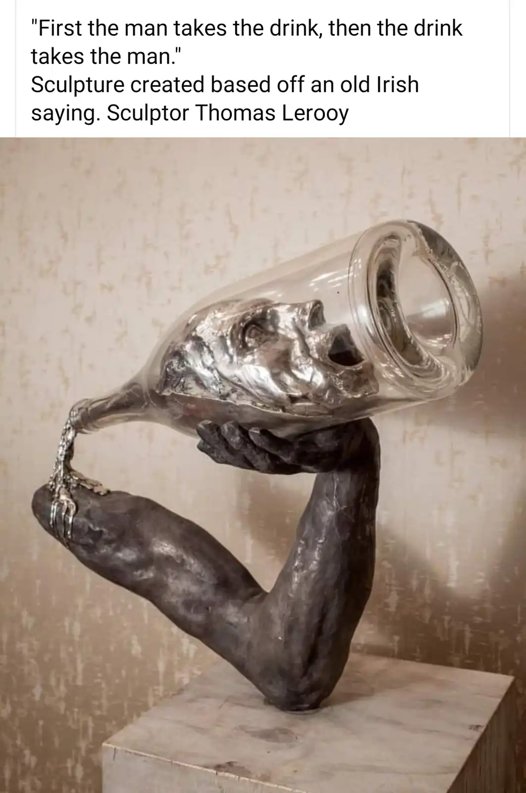 A sculpture depicts a muscular arm holding a large glass bottle, which is inverted and has a human face molded into its…