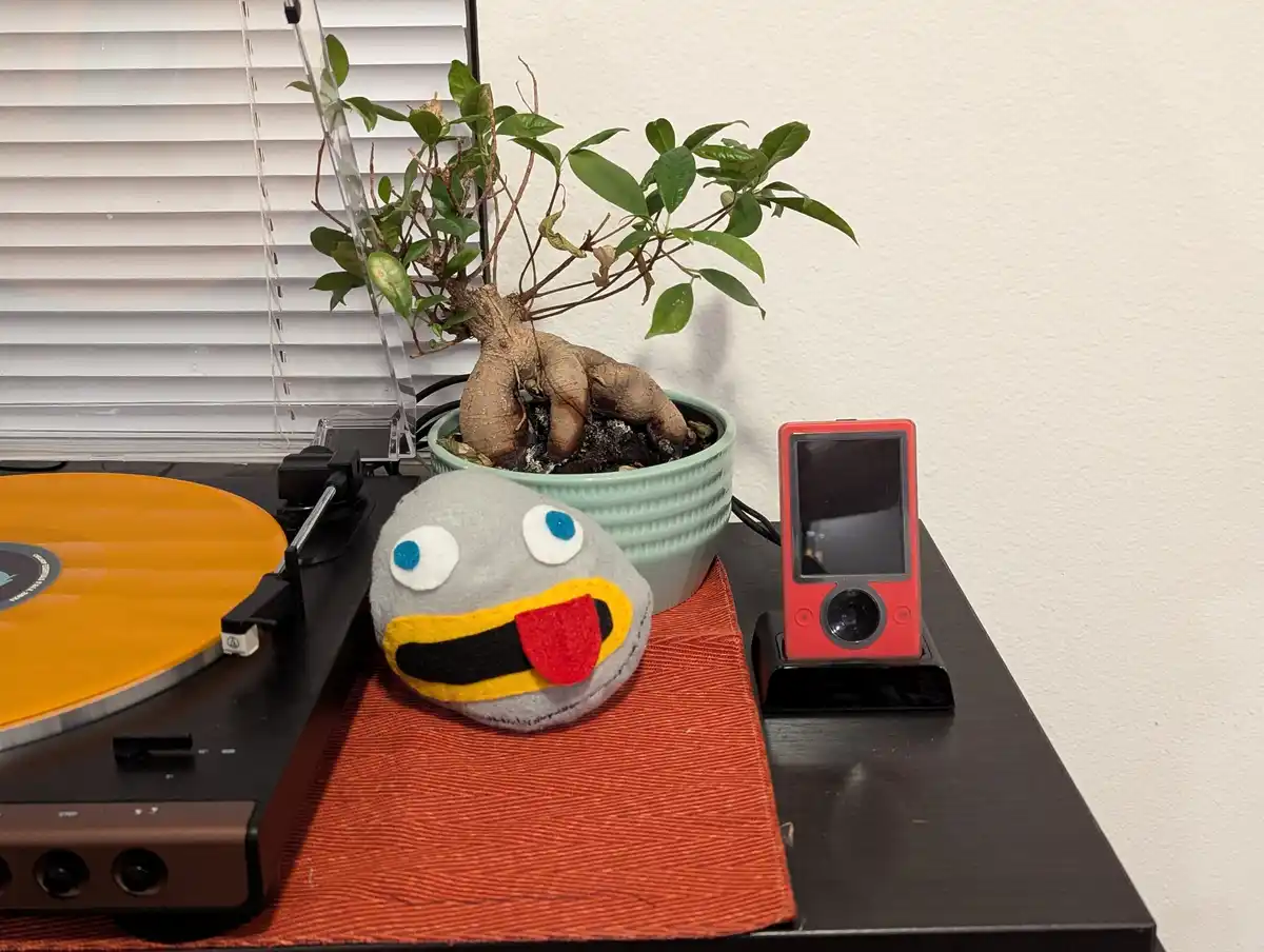 A felt plush toy with large blue eyes and a wide open mouth sits on a red mat next to a record player with an orange vinyl…