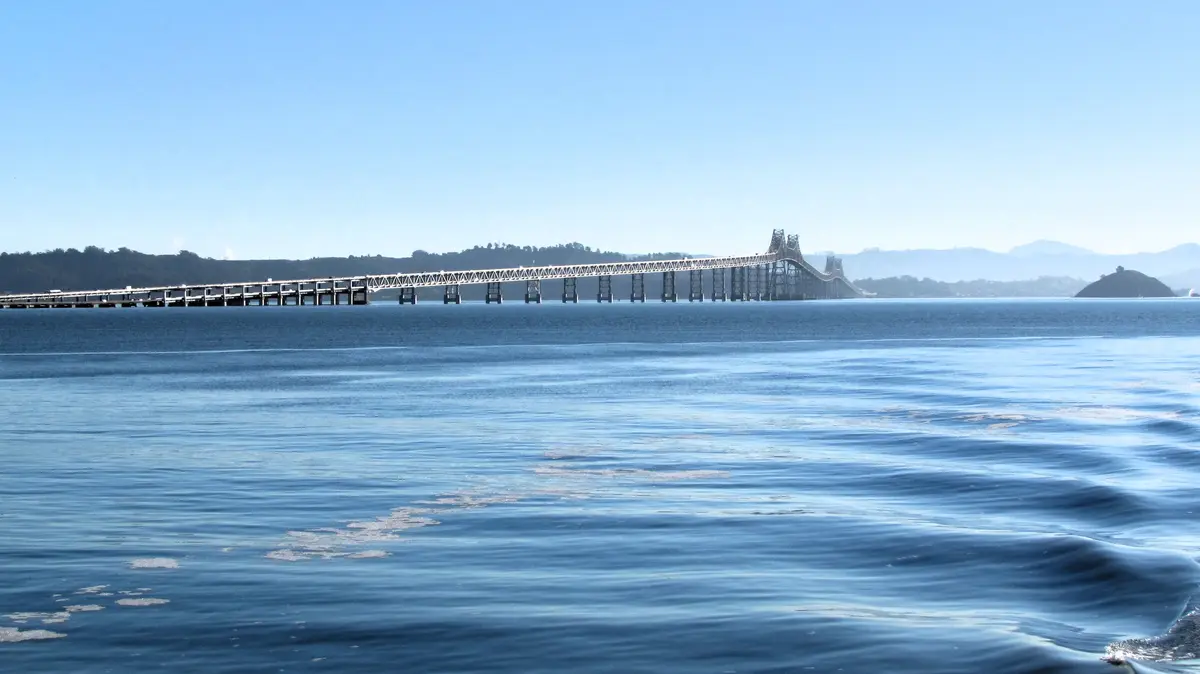A scenic photo of a long truss bridge spanning a wide body of blue water under a clear sky, with distant hills and a small…