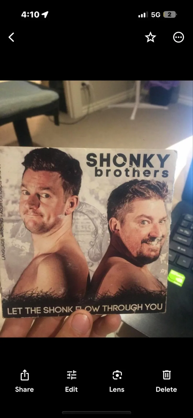 A person holds up a CD cover featuring two shirtless men with exaggerated facial expressions. The cover reads 'SHONKY…