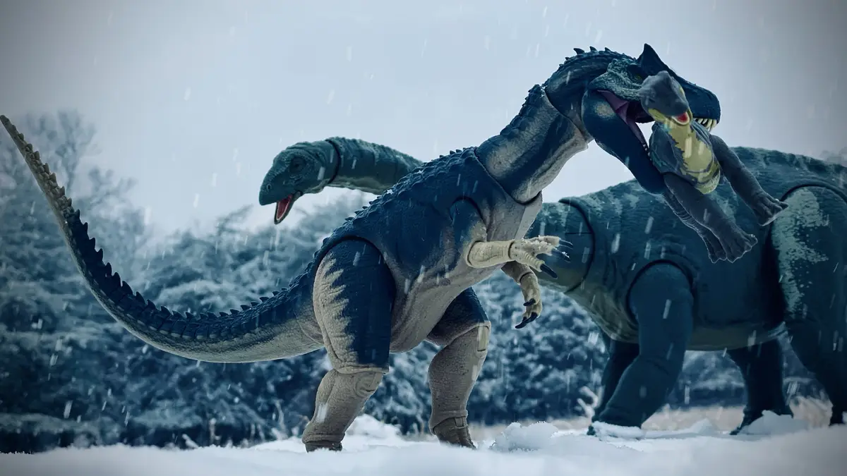 A blue toy dinosaur with a long tail and spines is standing in snow, holding a smaller dinosaur toy in its mouth. Another…