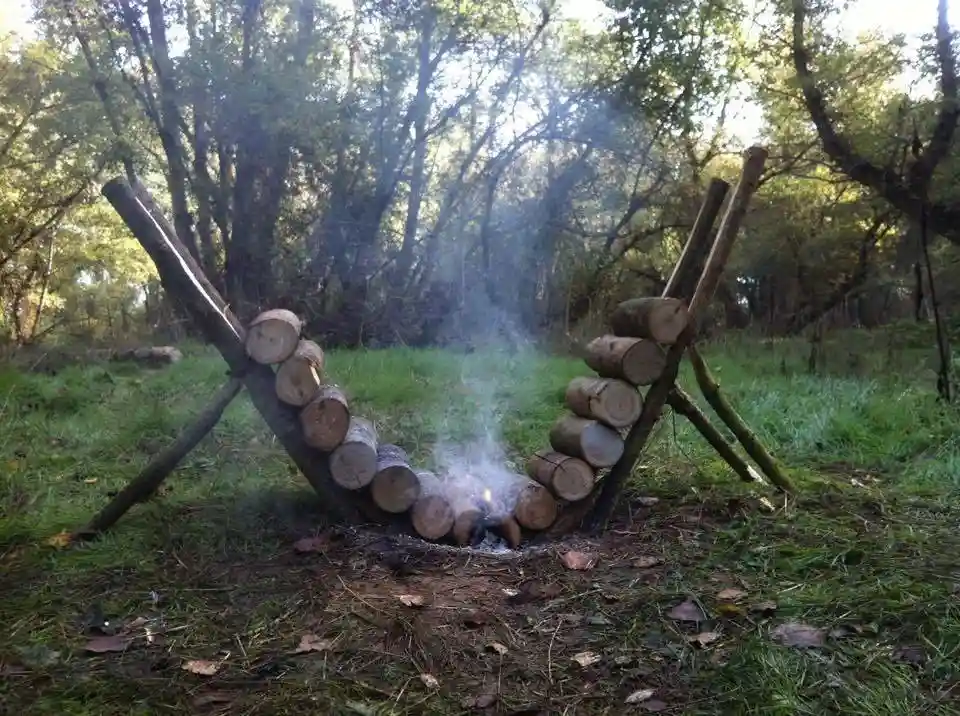 A rustic outdoor scene shows a fire pit constructed from logs arranged in a curved, hammock-like shape, with smoke rising…