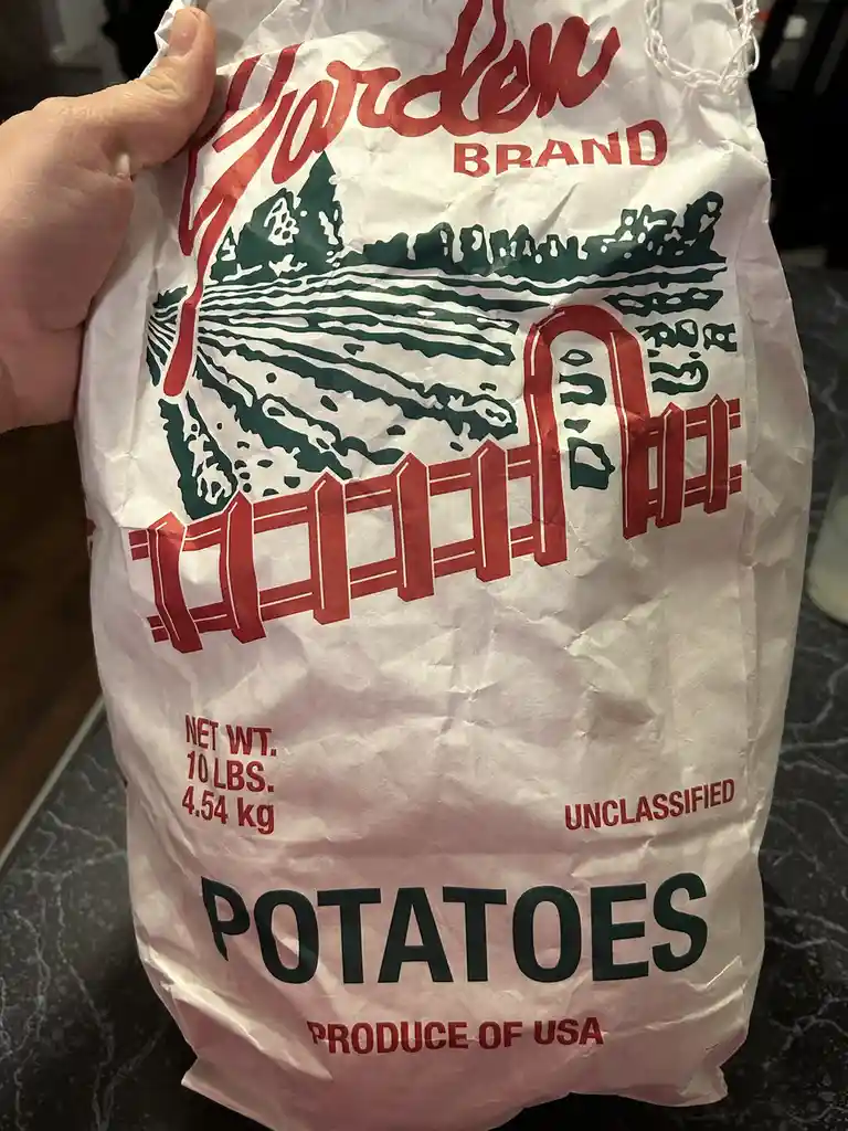 A hand holds a large, crinkled white paper bag of 'Garden Brand' potatoes, featuring a red and green logo with a fence…