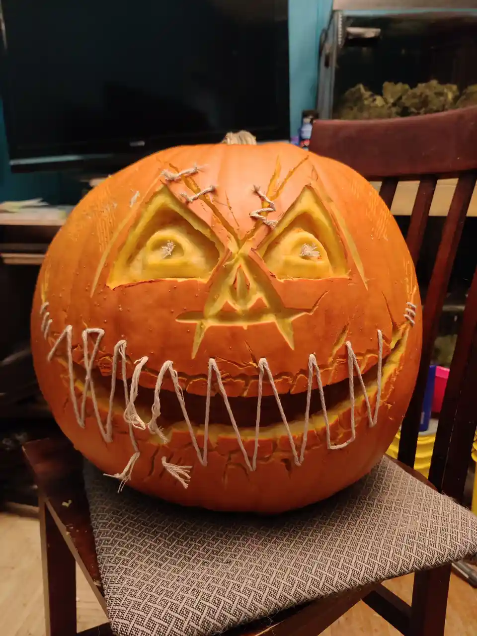 A carved orange pumpkin with a stitched-up, angry face sits on a wooden chair. The pumpkin has triangular eyes, a…