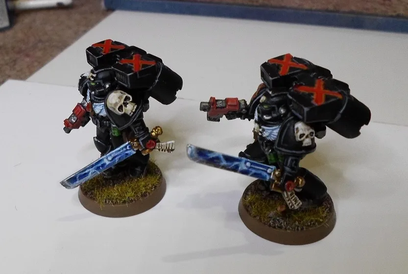 Two identical black-armored miniature figures with skull motifs and red cross symbols on their backs stand on grassy bases…
