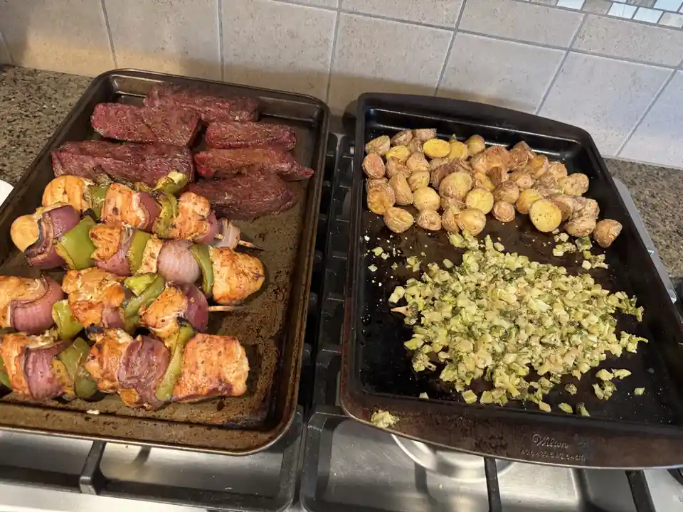 Two baking sheets sit on a stovetop; one holds large steak strips and chicken skewers with peppers and onions, the other…