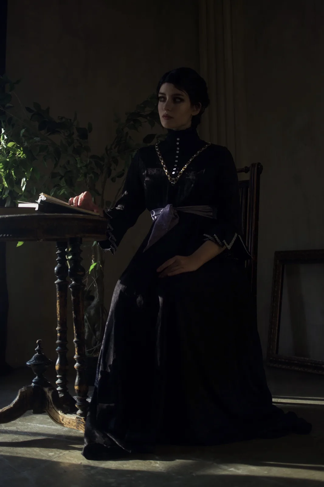 A woman in a dark, ornate Victorian-style dress sits at a wooden table in a dimly lit room, her face illuminated by a single…