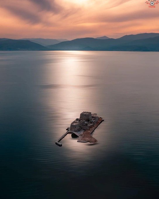 An aerial photo shows a dilapidated, partially submerged structure in calm water, with mountains and a sunset in the…