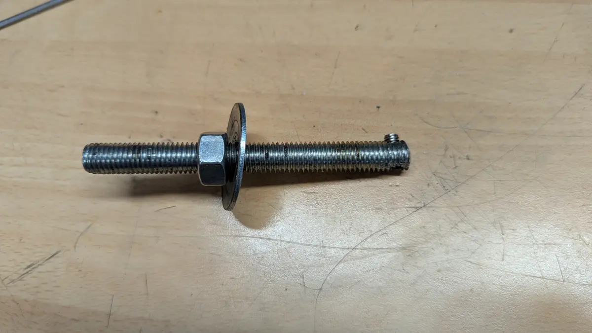 A metal bolt with a hex nut and washer lies horizontally on a scratched wooden surface. The bolt's head is missing, replaced…