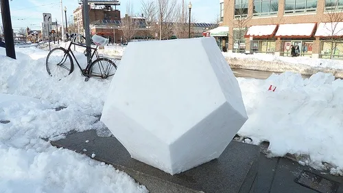 A large, geometric snow sculpture sits on a sidewalk next to a bicycle buried in snow, with a street and buildings in the…