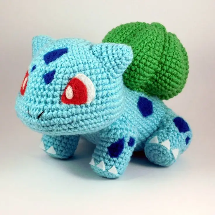 A handmade crocheted plush toy of the Pokémon Bulbasaur, featuring its signature blue body, green bulb on its back, red eyes…