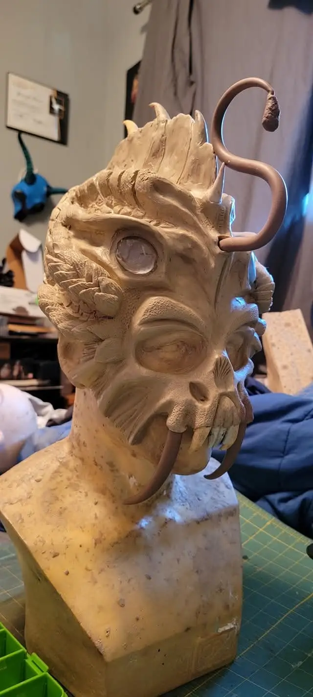 A clay sculpture of a demonic head with horns, tusks, and a curled tail-like appendage sits on a workbench. The text…