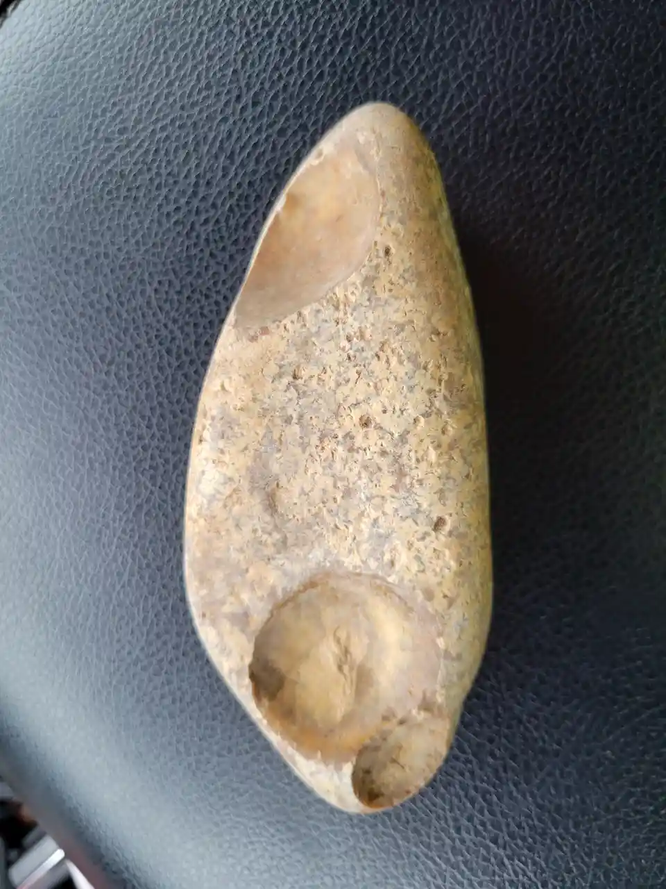 A smooth, tan-colored stone with a distinct elongated shape and a hollowed-out section at the bottom rests on a dark gray…