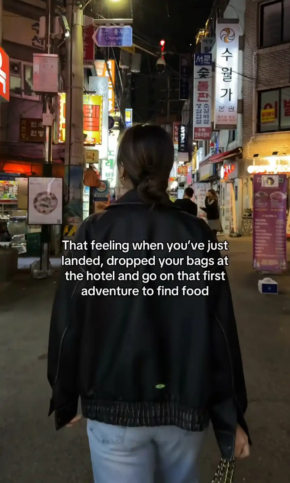 A person viewed from behind walks down a brightly lit, narrow Korean street at night, surrounded by glowing signs in Korean…