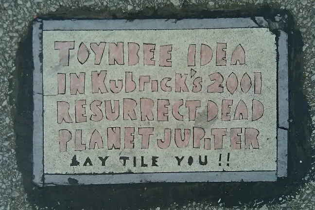 A close-up of a sidewalk tile with raised lettering that reads 'TOYNBEE IDEA IN Kubrick's 2001 RESURRECT DEAD PLANETJUPITER…