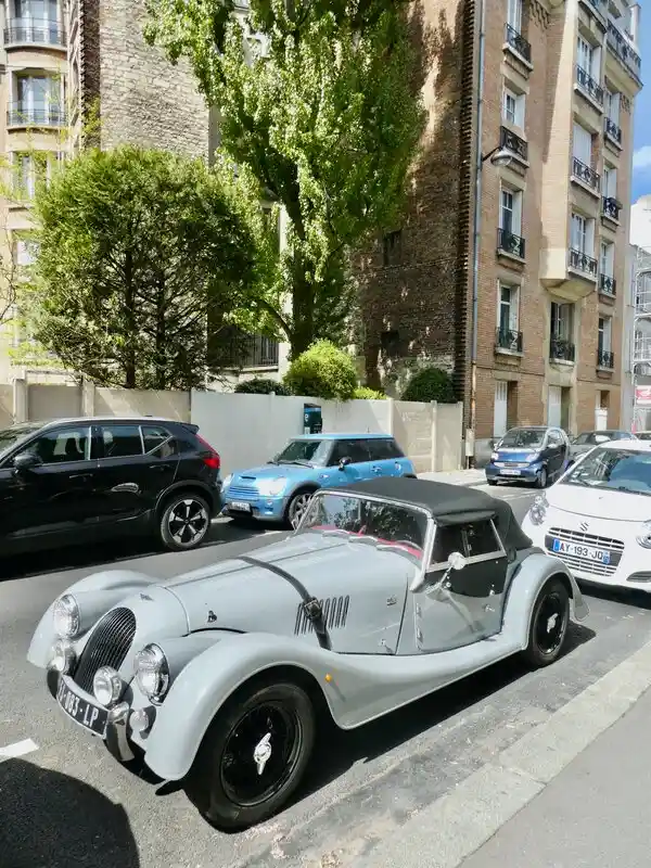 A light gray vintage Morgan roadster with a black soft top is parked on a city street, surrounded by modern cars including a…