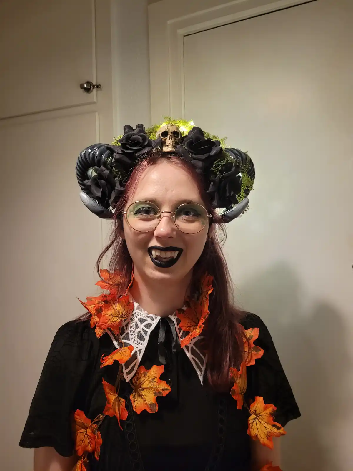 A person with red hair and glasses wears a Halloween costume featuring a black rose and skull crown with curved horns, black…