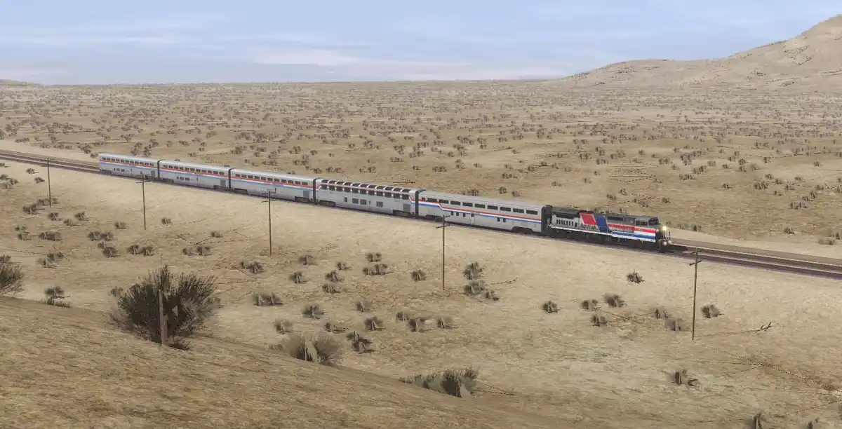 A long Amtrak passenger train with silver cars and red/white/blue stripes travels through a vast, arid desert landscape…