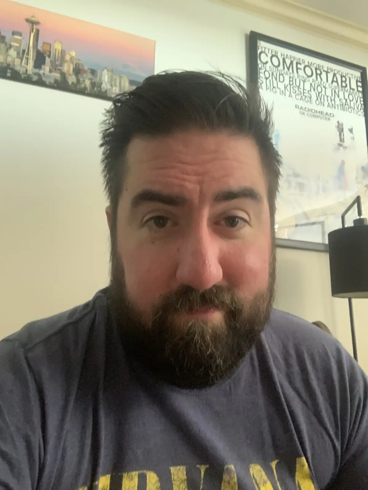 A man with a beard and dark hair stares directly at the camera with a weary, unimpressed expression. He wears a dark blue…