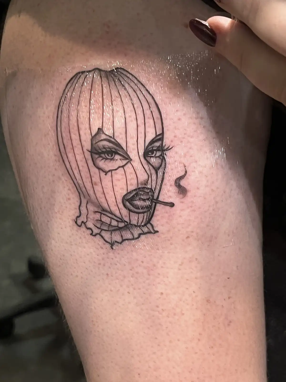 A freshly inked black-and-white tattoo on human skin shows a stylized woman's face wearing a balaclava with vertical lines…
