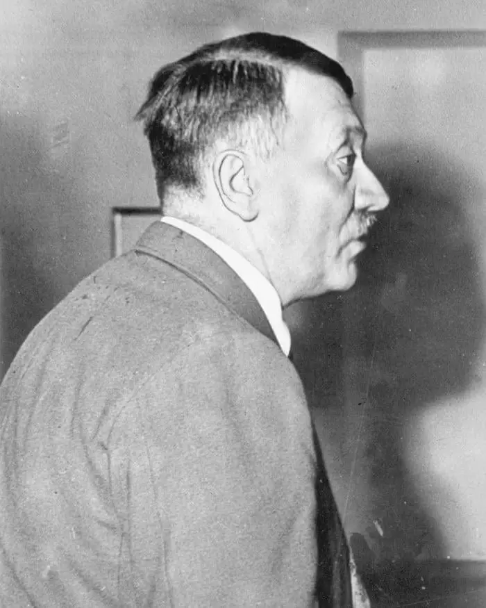A black-and-white historical photograph shows a man in profile, wearing a suit and tie, looking over his shoulder. The image…